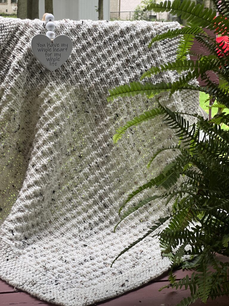 Ivory and tweed textured baby blanket on a red wood bench framed by a fern